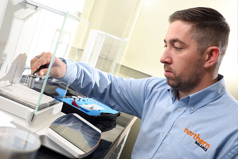 Man wearing a Northern Balance shirt is using tweezers to place a test weight on a micro balance